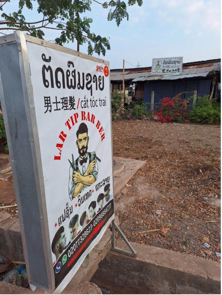 中国資本のサイセター総合開発区ゲート前の理髪店。 ラオス語、中国語とベトナム語の表記で「男性理髪」（2024年2月28日）
