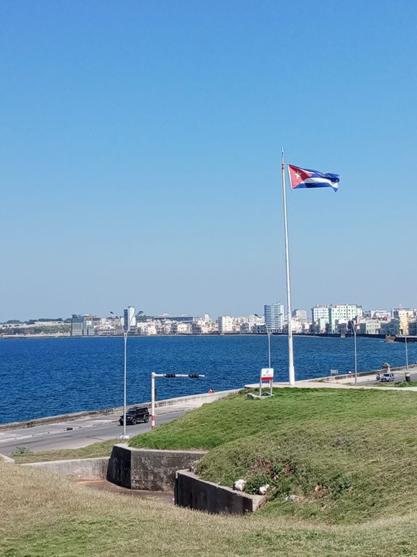 写真３：ハバナ市ベダド地区の海岸沿いにあるキューバを代表するホテル、ナシオナル（1930年建設）から。手前はキューバの国旗。ハバナ湾をはさんで向こう岸左半分、緑の見える部分に、スペインが建設した要塞の一つ、カバーニャ要塞が見える。（撮影：Sunamis Fabelo）